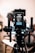 A professional camera setup mounted on a tripod dominates the foreground. The camera is equipped with various accessories, including a microphone with a windscreen, handles, and a visible battery pack labeled 'COMER D&O Lighting'. The background is softly blurred, showcasing the interior of a studio-like environment with a chair and monitor.