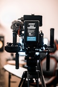 A professional camera setup mounted on a tripod dominates the foreground. The camera is equipped with various accessories, including a microphone with a windscreen, handles, and a visible battery pack labeled 'COMER D&O Lighting'. The background is softly blurred, showcasing the interior of a studio-like environment with a chair and monitor.