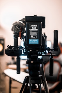 A professional camera setup mounted on a tripod dominates the foreground. The camera is equipped with various accessories, including a microphone with a windscreen, handles, and a visible battery pack labeled 'COMER D&O Lighting'. The background is softly blurred, showcasing the interior of a studio-like environment with a chair and monitor.