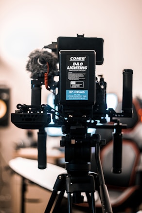 A professional camera setup mounted on a tripod dominates the foreground. The camera is equipped with various accessories, including a microphone with a windscreen, handles, and a visible battery pack labeled 'COMER D&O Lighting'. The background is softly blurred, showcasing the interior of a studio-like environment with a chair and monitor.