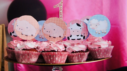 A group of cupcakes with pink frosting and polka dot wrappers, decorated with farm animal-themed toppers featuring pigs, cows, and sheep. A glittery number one is visible in the background, suggesting a first birthday celebration. The backdrop consists of shades of pink, with a black circular object partially visible.