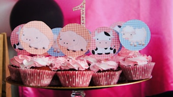 A group of cupcakes with pink frosting and polka dot wrappers, decorated with farm animal-themed toppers featuring pigs, cows, and sheep. A glittery number one is visible in the background, suggesting a first birthday celebration. The backdrop consists of shades of pink, with a black circular object partially visible.