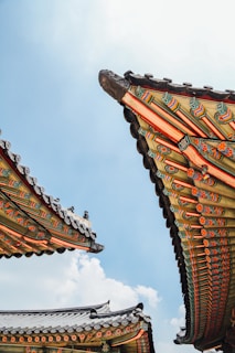 Intricate, colorful eaves of traditional Korean architecture against a bright blue sky. The ornate detailing includes floral and geometric patterns in vibrant shades of green, red, and gold.