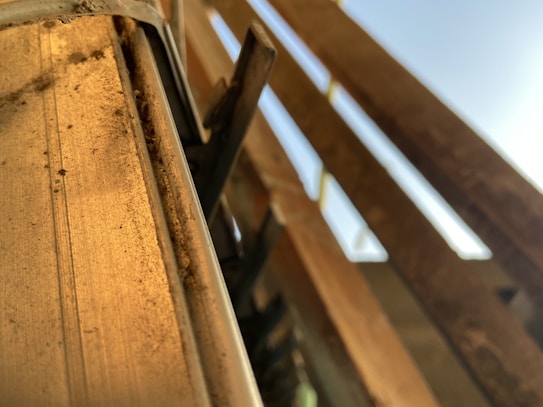 Close-up view of a rough, textured metal surface with a dusty appearance. Wooden beams or planks are visible to the right, casting shadows and reflecting light under a clear blue sky.