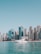A white yacht floats on calm turquoise waters with a backdrop of modern high-rise buildings under a clear blue sky. The skyline includes both glass and concrete structures of varying heights, suggesting an urban coastal scene.