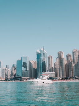 A white yacht floats on calm turquoise waters with a backdrop of modern high-rise buildings under a clear blue sky. The skyline includes both glass and concrete structures of varying heights, suggesting an urban coastal scene.