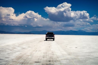 A pickup truck travels on a vast, flat expanse of white salt flats under a sky filled with large, dramatic clouds. In the background, distant mountains are visible along the horizon.