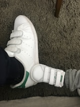 A pair of white sneakers with green accents and Velcro straps is seen on a shaggy grey carpet. One shoe is significantly larger than the other, suggesting the presence of an adult and a child.