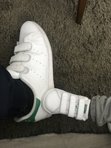 A pair of white sneakers with green accents and Velcro straps is seen on a shaggy grey carpet. One shoe is significantly larger than the other, suggesting the presence of an adult and a child.