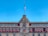 A historic building with an ornate central facade and numerous windows under decorative arches. The building features a large Mexican flag atop a flagpole, and red awnings cover the windows on the lower level. The architecture combines intricate stone carvings with a symmetrical design.