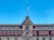 A historic building with an ornate central facade and numerous windows under decorative arches. The building features a large Mexican flag atop a flagpole, and red awnings cover the windows on the lower level. The architecture combines intricate stone carvings with a symmetrical design.