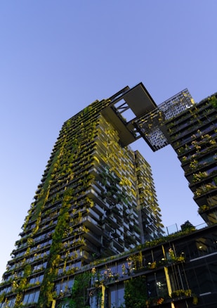 A tall modern building covered in lush greenery and plants, with a unique architectural design featuring a large sky garden or terrace near the top. The building stands against a clear blue sky, and the sunlight illuminates the plants, creating a vibrant and natural facade.