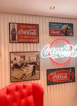 A vintage-style interior display features various Coca-Cola advertisements. On the wall, framed posters include images of a classic Coca-Cola bottle and nostalgic artwork depicting people enjoying the beverage. The centerpiece is a bright neon Coca-Cola sign. A plush red chair is partially visible in the foreground, adding to the retro atmosphere.