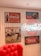 A vintage-style interior display features various Coca-Cola advertisements. On the wall, framed posters include images of a classic Coca-Cola bottle and nostalgic artwork depicting people enjoying the beverage. The centerpiece is a bright neon Coca-Cola sign. A plush red chair is partially visible in the foreground, adding to the retro atmosphere.