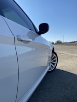 The photo captures the front side of a white car parked on a paved surface. The sun reflects off the smooth surface of the car, highlighting its sleek design. A vast expanse of clear blue sky stretches overhead, with a faint silhouette of distant trees and a solitary utility pole visible in the background.