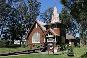 A quaint wooden chapel with a spire sits amidst a lush green landscape. The chapel features a steep, gabled roof, wood siding, and a ramp leading to the entrance. Surrounded by tall trees and flowering shrubs, this serene setting is complemented by bright daylight.