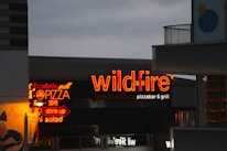 The scene features brightly illuminated neon signs advertising a pizza bar and grill with the name 'wildfire' in bold red letters. One of the signs is written in both English and possibly another language, highlighting menu items like pizza, salad, and drinks. The surrounding buildings are partially visible, with a cloudy evening sky providing a contrasting backdrop.