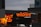 The scene features brightly illuminated neon signs advertising a pizza bar and grill with the name 'wildfire' in bold red letters. One of the signs is written in both English and possibly another language, highlighting menu items like pizza, salad, and drinks. The surrounding buildings are partially visible, with a cloudy evening sky providing a contrasting backdrop.