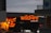 The scene features brightly illuminated neon signs advertising a pizza bar and grill with the name 'wildfire' in bold red letters. One of the signs is written in both English and possibly another language, highlighting menu items like pizza, salad, and drinks. The surrounding buildings are partially visible, with a cloudy evening sky providing a contrasting backdrop.