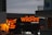The scene features brightly illuminated neon signs advertising a pizza bar and grill with the name 'wildfire' in bold red letters. One of the signs is written in both English and possibly another language, highlighting menu items like pizza, salad, and drinks. The surrounding buildings are partially visible, with a cloudy evening sky providing a contrasting backdrop.