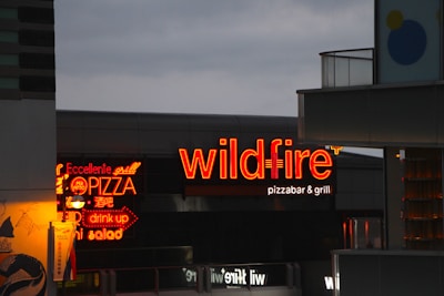 The scene features brightly illuminated neon signs advertising a pizza bar and grill with the name 'wildfire' in bold red letters. One of the signs is written in both English and possibly another language, highlighting menu items like pizza, salad, and drinks. The surrounding buildings are partially visible, with a cloudy evening sky providing a contrasting backdrop.