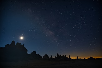 A starry night sky with a bright celestial body, possibly the moon or a bright planet, illuminating the dark silhouette of jagged rock formations. The horizon shows a faint glow, suggesting a transition from night to dawn.