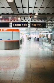 A modern airport terminal with a sleek design, featuring a tiled floor and metallic ceiling. Overhead signs indicate directions and services such as restrooms, baggage claim, and cafes. A travel information desk with a bright red-orange counter is positioned on the left, while the area appears spacious with some people in the background.
