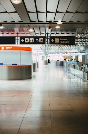 A modern airport terminal with a sleek design, featuring a tiled floor and metallic ceiling. Overhead signs indicate directions and services such as restrooms, baggage claim, and cafes. A travel information desk with a bright red-orange counter is positioned on the left, while the area appears spacious with some people in the background.