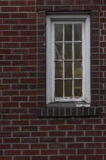 A weathered, narrow window with multiple panes set in a red brick wall. The white paint on the window frame is chipped, and the glass appears slightly foggy.