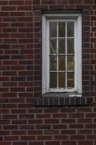 A weathered, narrow window with multiple panes set in a red brick wall. The white paint on the window frame is chipped, and the glass appears slightly foggy.