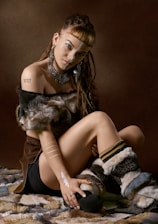 A person with braided hair sits on a fur-covered surface. They have various body adornments, including large earrings, a necklace, nose jewelry, and body paint. The person is wearing fur-like boots and a top with fur trim, and has visible tattoos on their arm and chest.