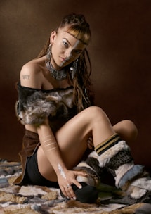A person with braided hair sits on a fur-covered surface. They have various body adornments, including large earrings, a necklace, nose jewelry, and body paint. The person is wearing fur-like boots and a top with fur trim, and has visible tattoos on their arm and chest.