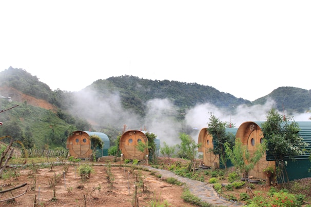 A picturesque landscape featuring several barrel-shaped wooden cabins with green roofs, set amidst a lush, mountainous area. Light mist or fog rises around the cabins, suggesting a cool, serene atmosphere. The foreground shows a small garden with young plants and a stone path leading towards the cabins.
