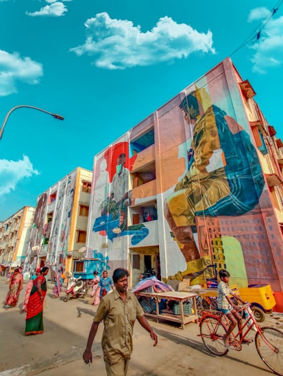 A vibrant street scene featuring a building adorned with large, colorful murals depicting people engaged in various activities. The street is lively with pedestrians, including women in traditional attire and a man walking past, while a child rides a red bicycle. A clear blue sky with scattered clouds provides a bright contrast to the vivid colors of the murals.