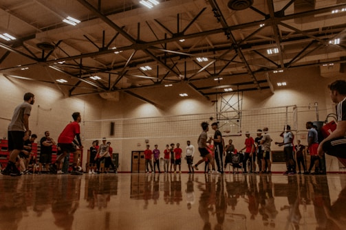 A group of people engaged in a volleyball game inside a well-lit gymnasium. Several participants are lined up along the sides of the court, watching and waiting for their turn to play. The setting features polished wooden flooring, and the ceiling is equipped with industrial-style lighting fixtures.
