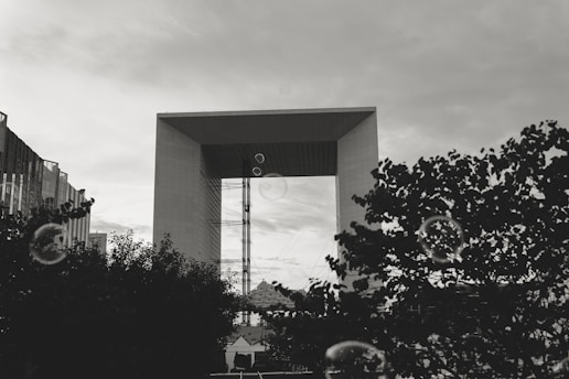 A large, modern arch-shaped building stands prominently against a cloudy sky. In the foreground, silhouettes of tree branches create a contrasting frame. Several floating bubbles add an interesting and playful element to the scene.