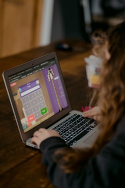 A person using a laptop on a wooden table, engaged in what appears to be a math activity on an interactive website. The screen displays a calculator interface with numbers and operational buttons. The person's focus is directed towards the screen.