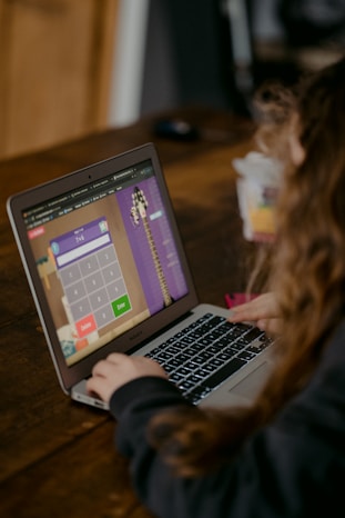 A person using a laptop on a wooden table, engaged in what appears to be a math activity on an interactive website. The screen displays a calculator interface with numbers and operational buttons. The person's focus is directed towards the screen.