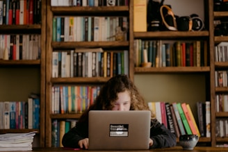 A young girl with curly hair focuses intently on a laptop in a cozy room filled with bookshelves. The shelves are packed with a variety of colorful books and small decorative items. A cup sits on the wooden table beside her.