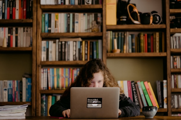 A young girl with curly hair focuses intently on a laptop in a cozy room filled with bookshelves. The shelves are packed with a variety of colorful books and small decorative items. A cup sits on the wooden table beside her.
