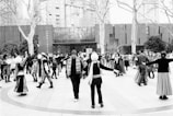 A large group of people gathers in an open outdoor space, engaging in what appears to be a community dance or social gathering. Many participants are dressed in traditional or cultural attire, with some wearing hats and scarves. The background shows a modern building surrounded by trees and shrubs.