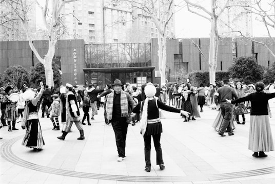 A large group of people gathers in an open outdoor space, engaging in what appears to be a community dance or social gathering. Many participants are dressed in traditional or cultural attire, with some wearing hats and scarves. The background shows a modern building surrounded by trees and shrubs.