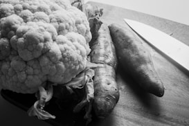 A large, textured head of cauliflower and two carrots rest on a wooden cutting board. A sharp kitchen knife lies nearby, suggesting preparation for cooking. The image's monochromatic tone adds a classic and timeless feel.