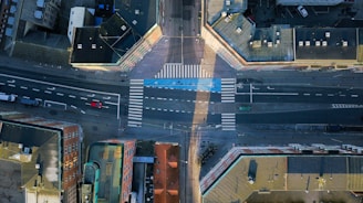 An aerial view of an urban intersection with multiple buildings surrounding the roads. The streets are marked with white lines and crosswalks, including distinct bicycle lanes highlighted in blue. A few vehicles can be seen on the roads, including a red car and a white truck.