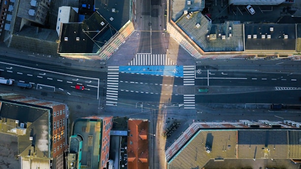 An aerial view of an urban intersection with multiple buildings surrounding the roads. The streets are marked with white lines and crosswalks, including distinct bicycle lanes highlighted in blue. A few vehicles can be seen on the roads, including a red car and a white truck.