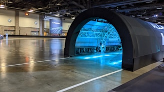 A large indoor space that resembles a convention center or exhibition hall. There is a black arched structure, possibly an entrance or tunnel, illuminated with blue lights from within. The flooring is reflective and the space is relatively empty. There are some signs and doors on the walls, and in the background, a large banner with the text 'I-PACE' is visible.