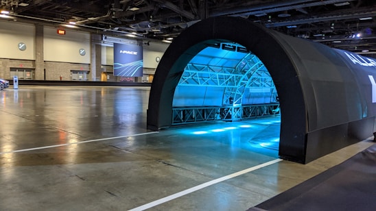 A large indoor space that resembles a convention center or exhibition hall. There is a black arched structure, possibly an entrance or tunnel, illuminated with blue lights from within. The flooring is reflective and the space is relatively empty. There are some signs and doors on the walls, and in the background, a large banner with the text 'I-PACE' is visible.