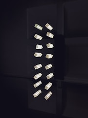 A collection of geometric light fixtures oriented vertically in a dark setting. The lights are rectangular with a luminous quality, emitting a soft glow against the shadowy backdrop. The fixtures are arranged in a seemingly random yet organized pattern, creating a minimalist and modern aesthetic.