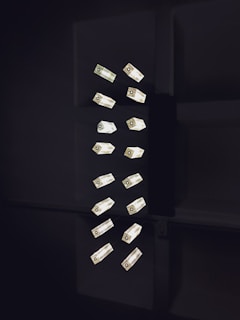 A collection of geometric light fixtures oriented vertically in a dark setting. The lights are rectangular with a luminous quality, emitting a soft glow against the shadowy backdrop. The fixtures are arranged in a seemingly random yet organized pattern, creating a minimalist and modern aesthetic.