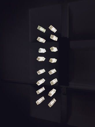A collection of geometric light fixtures oriented vertically in a dark setting. The lights are rectangular with a luminous quality, emitting a soft glow against the shadowy backdrop. The fixtures are arranged in a seemingly random yet organized pattern, creating a minimalist and modern aesthetic.